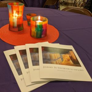 close up of a table covered with a purple tablecloth, holding three "stained glass" style tea light candles next to several service programs featuring a painting of a lighted street in NT Bethlehem, surrounded by a dark sky.