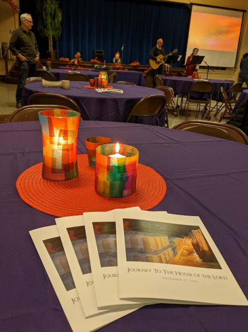 A church basement with musicians and a projection screen in the background, preparing for the Sunday service; close up of a table covered with a purple tablecloth, holding three "stained glass" style tea light candles next to several service programs featuring a painting of a lighted street in NT Bethlehem, surrounded by a dark sky.